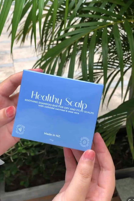Hand holding a blue box labeled 'Healthy Scalp' with green foliage in the background